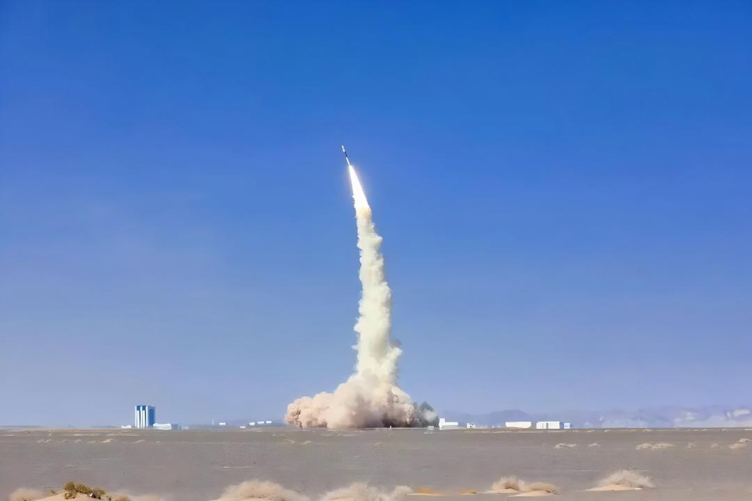Rocket climbing above a ground launch site with a long white exhaust trail.