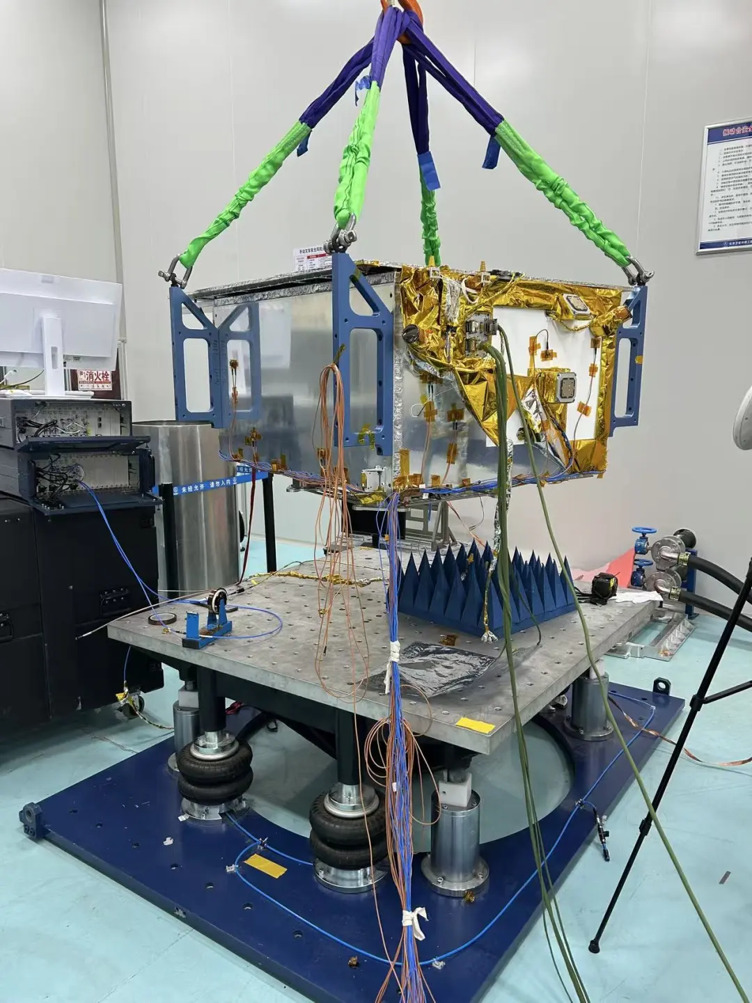Satellite assembly suspended by lifting straps above laboratory test equipment.