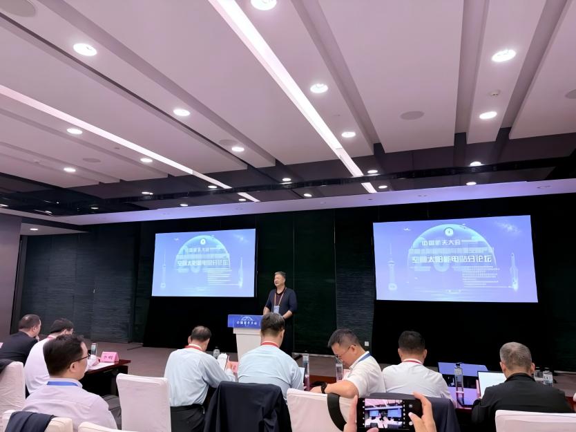 Speaker addressing a seated audience in a conference room with presentation screens.